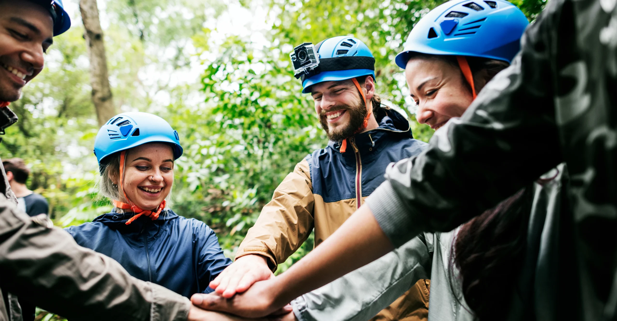 Glade mennesker i blå hjelme stabler hænder til teambuilding i naturen.